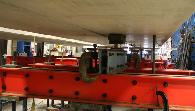 Figure 5. Clamping at the end of beams used in partially and fully stiffened cases. This photo highlights the clamping system that is used to reduce torsional rotation at the beam ends. The clamping system is highlighted in the photograph within a dashed rectangle. Two double channels are shown spanning transversely across the end of the beams. These steel channels serve to clamp down on the beam ends, attaching them to the strong floor through the use of threaded rods.Figure 5. Photo. Intermediate support on one beam used in the fully stiffened case. This photo highlights the in-span supports that reduce in-span deflections of one beam. This consists of a neoprene support near the connection between the two beams and a pulldown force applied by a threaded rod toward the outside of the same beam. The other beam, shown to the left, is not supported or held down at this location.