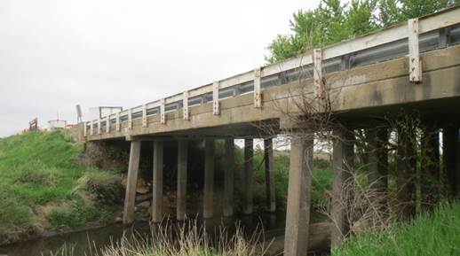 Figure 14. This figure shows a photo of the Laporte Road bridge from the elevation view. All three spans of the reinforced concrete slab bridge are shown. The bridge is supported at the ends by reinforced concrete abutments that have been cast over steel H-piles. The abutment-to-abutment length of the bridge is 100 ft (30.5 m). The width of the bridge is 28 ft (8.53 m) and services two lanes of traffic. The interior spans of the bridge are supported by H-piles that were permanently encased on concrete. The reinforced concrete slab superstructure of the bridge is composed of three spans that measure 30.5 ft (9.3 m), 39 ft (11.9 m), and 30.5 ft (9.3 m). The slab has a uniform thickness of 16.9 in (430 mm). The bridge deck has a 5 percent super elevation.