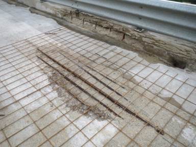 Figure 16. This figure shows a photo of the distressed region location on the westbound lane over pier 1. A large, oval-shaped region of distressed concrete can be observed. Within the oval-shaped region, five corroded steel reinforcing bars are present. The photo also shows that there is some deterioration present along the curb of the bridge structure.