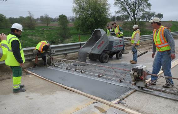 Figure 19. This photo shows the UHPC overlay material being placed on the westbound lane of the bridge. This was the first of two installation phases. UHPC is being placed using a motorized buggy. Workers are assisting with placement using hand tools. The overlay is being finished using a large vibrating screed that the workers move manually.