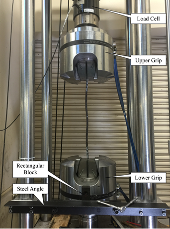 In this view of the load frame, the four chromed columns are running vertically on the left and right sides of the photo. At the bottom of the photo is a black steel angle oriented horizontally and bolted between the left and right front chromed columns. Behind the steel angle is the lower hydraulic grip, and to the front of the lower grip is a black vertically oriented steel block that is bolted to the grip. At the top center of the photo is the load cell, and connected to the load cell is the upper hydraulic grip.