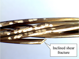 This is a closeup photo of an unsheathed strand. The wires are helically wrapped, so there is a slight angle from lower left to upper right. The wires are noticeably separated from each other. There is a broken wire at the bottom; its fracture surface is diagonally inclined relative to the longitudinal direction of the wire. An annotation points to the fracture surface called out as &ldquo;Inclined shear fracture.&rdquo;