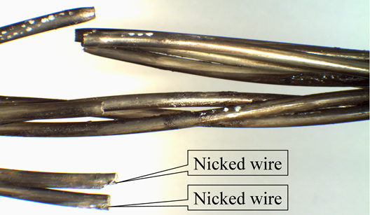 This is a closeup view of a strand where four wires have fractured. Due to the fracture, the wires are splayed in multiple directions. The two fractured wires at the bottom of the photo show that, at the fracture location, the fracture appears to have initiated from a gouge in the wire. An annotation with a callout of &ldquo;Nicked wire&rdquo; points to each of these locations.