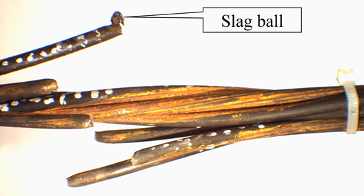 This is a closeup view of a strand where three wires have fractured. Due to the fracture, the wires are splayed upward on the left and downward on the right. The uppermost fractured wire has a small ball of metal fused to the surface of the wire directly at the fracture. An annotation with a callout of &ldquo;Slag ball&rdquo; points to this location.