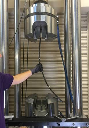 In this view of the load frame, the four chromed columns are running vertically on the left and right sides of the photo. At the bottom of the photo is a black steel angle oriented horizontally and bolted between the left and right chromed columns. Behind the steel angle is the lower hydraulic grip. At the top center of the photo is the load cell and upper hydraulic grip. A hand is shown holding a strand before being gripped. The strand is centered on the upper grip but shown sweeping to the right so the other end is about 1 ft away from the center of the lower grip.