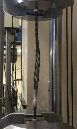 In this closeup view of the load frame, only a portion of the upper grip is visible at the top of the photo and only a portion of the lower grip at the bottom of the photo. A strand is gripped between the upper and lower grips. The strand is an obvious birdcage where all outer wires are clearly unwound from the king wires and spaced well apart from each other. The strand is also not straight; it has slight double curvature along its length where it deviates from the centerline of the grips by about one strand diameter.