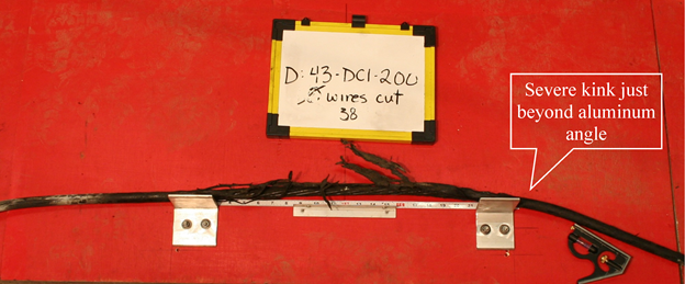 This photo shows strand D38 laying atop the measuring jig. The strand rests nearly flat upon the angles of the measuring jig; however, just to the right of the right-handed angle, the strand sharply bends downward. An annotation points to this location with the wording &ldquo;Severe kink just beyond aluminum angle.&rdquo;