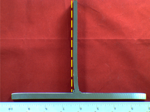 This photo shows a steel, T-shaped cross section. The flange is running horizontally across the bottom of the figure parallel to a ruler. The scale of the ruler is not shown, but the width of the flange is about 8 inches long. The web is vertically oriented and joined at the midwidth of the flange. A straight, dashed vertical line is shown to the left side of the web. The web is parallel to the line except for the lower one unit of length near the joint between the web and flange. It is clear the web suffered a horizontal offset to the left during the welding event.