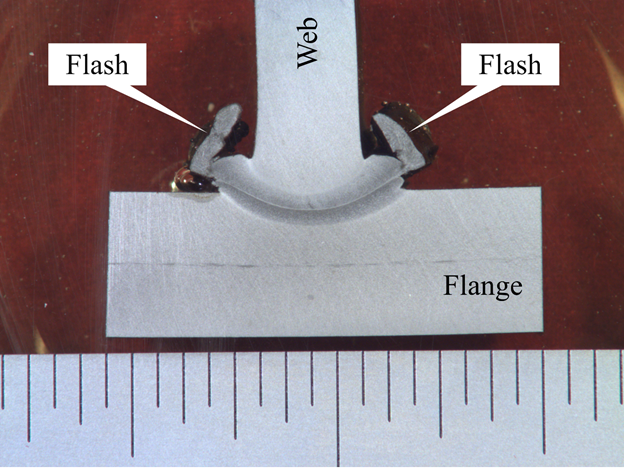This photo shows a macroetch of a small section of web, flange, and the complete joint penetration weld between the two. The flange plate (labeled “Flange”) is oriented horizontally, and the web (labeled “Web”) is vertical. The weld is arc shaped with a low point approximately one-tenth of a web thickness into the flange. A narrow line can be seen through the middle of the weld defining the fusion line, and on each side of the fusion line is a darker heat-affected zone. An irregular-sized piece of metal is shown outside of the joint on each side of the web and is labeled “Flash.” A ruler is provided in the field of view with uniform divisions, though units are not shown. The flange portion is 17 units wide, and by inspection, the web is 8 units high.
