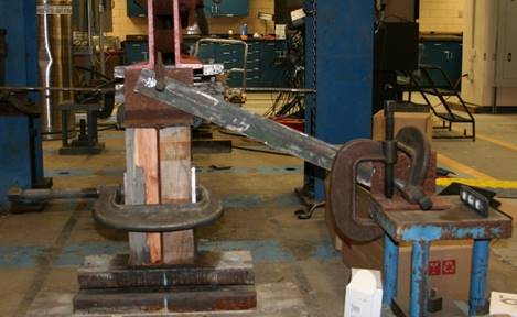 This photo shows an end view of the beam cross section on the left side. On each side of the web are wooden blocks fit between the inner surfaces of the flanges. A large C-clamp is clamping the two wooden blocks together with the web in the middle. An angle section is clamped to the top of the beam flange, and it has an outstanding leg upward away from the top flange. A pedestal is shown on the right side of the photo acting as a rigid support. An angle section is clamped to the top of the pedestal, and it has an outstanding leg upward away from the top of the pedestal. Another angle has been clamped to the outstanding legs of angles clamped to the top flange and to the pedestal. This angle slopes downward from the beam on the left to the pedestal on the right.