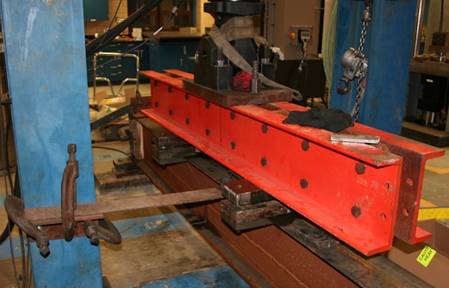 This photo shows a blue column of the load frame on the left side. An angle section is clamped to the flange of the column with two C-clamps such that there is an outstanding leg of angle that is parallel to the strong floor. The beam fatigue specimen runs from the lower right corner of the photo into the foreground at the upper middle of the photo. A thin piece of metal is clamped to the top flange of the beam fatigue specimen, oriented parallel to the strong floor and C-clamped to the outstanding leg of angle attached to the column. 