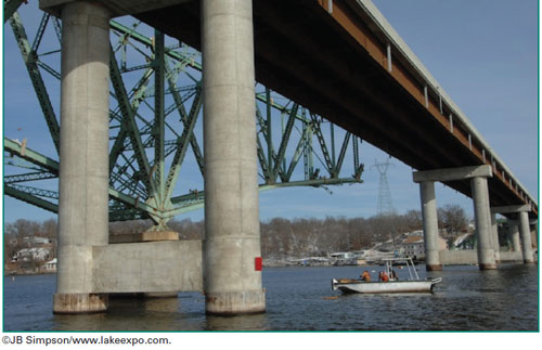 Figure 6. Photo. Hurricane Deck Bridge, Lake of the Ozarks, MO. The figure is a photo of the Hurricane Deck Bridge over the Lake of the Ozarks, MO. The photo is taken at a long distance from the bridge, with the photographer probably sitting in a boat. The photo shows about 3.5 spans of the new, concrete Hurricane Deck Bridge crossing the river. The photo shows four pier lines over the river. Each pier line consists of a pair of concrete columns connected at the bottom just above the water line and on top by the pier cap. Next to the near pier line, a steel arch truss bridge is shown that is cut off (demolished) about halfway through the next column line. The pier of the old truss bridge is close to the near pier line of the new bridge, with a small boat shown passing between the two piers. The distance between the old and new piers appears to be about 1 boat length.