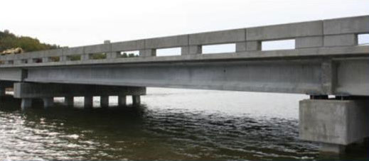The photograph shows one span of the bridge over water. The side of an exterior girder, the side of the bridge deck, and the side of the barrier railing are visible, along with the pier cap that supports the ends of this span.
