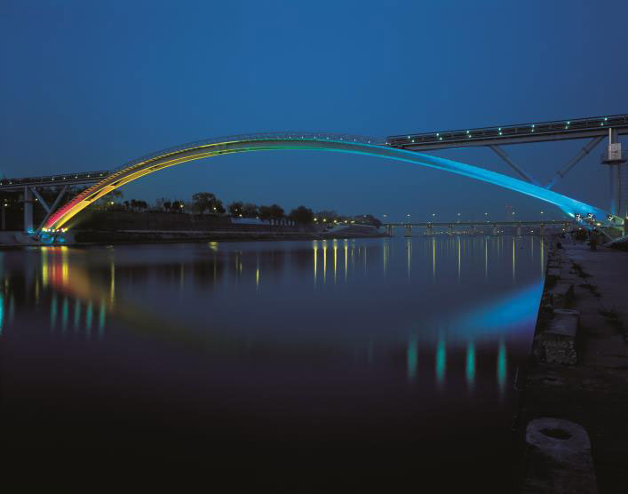 The photograph shows an elevation of the arch bridge over water taken at night from the river bank.