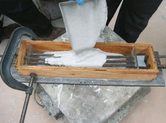 This figure shows the placement of ultra-high performance concrete (UHPC) into the formwork for an individual test specimen. The three strands are visible as the UHPC is deposited into the form. 