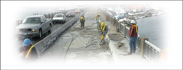 Workmen using jackhammers on a closed lane on a bridge with traffic moving by in a single lane beside them.