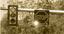 This photograph shows a traffic signal hanging on a horizontal pole over a roadway. An illuminated stop sign is shown hanging next to the signal. 