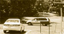 This photograph shows one car approaching an intersection and another car in the middle of the intersection. Two traffic signals and an illuminated stop sign are shown hanging above the intersection to control the traffic as it moves through the intersection.