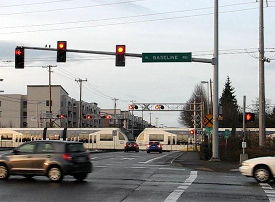 This photo shows a signalized intersection located in advance of a rail crossing with a light rail train traversing the crossing. The signal is red, and the crossing is closed to traffic, with queued vehicles waiting to proceed over the crossing.