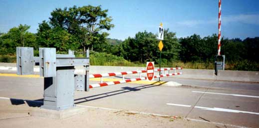 This photo shows a real-world implementation of an automated gate, which blocks access to a section of roadway. In the background of the roadway, a rural terrain with tall trees and blue skies is visible. Red-and-white-striped metal bars across the roadway are similar to what is used at railroad crossings. A typical “DO NOT ENTER” sign is located in the middle of these horizontal metal bars. The roadway has two lanes in each direction, with a small concrete median between directions. In one direction of travel, the automated gate is in the down position, blocking two lanes. In the other direction of travel, the automated gate is in the up position, allowing access to two lanes.
