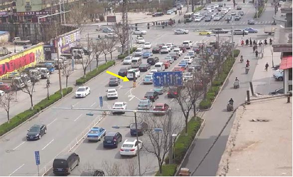 This photo focuses on the northbound approach to a signalized intersection in China. In the middle of this photo is a presignal that controls entry to two contraflow left-turn (CLT) pockets. Approximately 600 ft of the northbound approach lanes are visible within the photo. A queue of nine left-turning vehicles extends back to a presignal location, approximately 300 ft upstream of the main intersection. The presignal display is green, with a yellow arrow pointing to it, which implies that left-turning vehicles are allowed to enter the CLT pockets during this portion of the signal cycle. Twelve vehicles have entered the CLT pockets, and are beginning to queue up at the main intersection. Additional vehicles are entering the CLT pockets from the presignal location.