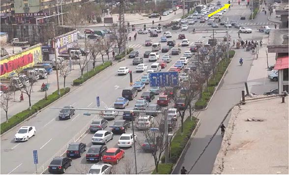 This photo focuses on the northbound approach to a signalized intersection in China. In the middle of this photo is a presignal that controls entry to two contraflow left-turn (CLT) pockets. Approximately 600 ft of the northbound approach lanes are visible within the photo. The queue of left-turning vehicles is so long that it extends beyond the bottom edge of the photo (i.e., beyond 600 ft). There are 17 vehicles visibly queued up in the left-turn lane before reaching the bottom edge of the photo. The presignal is located in the middle of the visible queue length (i.e., approximately 300 ft upstream of the main intersection), and in the median between both directions of travel. The presignal display is red, with a yellow arrow pointing to it, which implies that left-turning vehicles are not allowed to enter the CLT pockets during this portion of the signal cycle. However northbound left-turning vehicles have begun moving through the main intersection from three separate lanes (i.e., one conventional left-turn lane and two CLT pockets). The photo clearly shows that the availability of CLT pockets increases the number of left-turning vehicles that are able to move through this intersection.