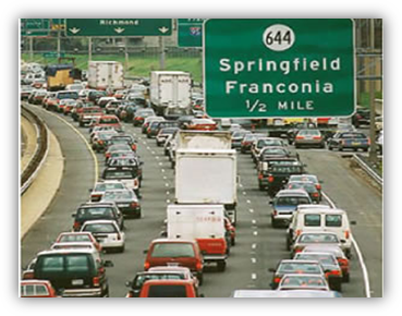 Image shows heavy traffic traveling on a three-lane highway. Traffic is a mix of personal cars and tractor trailers. There are orange cones along the road, indicating roadwork.