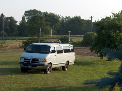 Figure 8. Photo. Antenna configuration on van used for collecting data broadcast from HAG1 and HRN2 simultaneously. The van is equipped with one GPS marine antenna and two demodulator receiver antennas.  Picture of a large white van with several antennas mounted on top.  The van is facing toward the lower left hand corner of the photo. On the left side of the van toward the front is a GPS antenna; toward the back is a second GPS antenna.  On the right side of the van are mounted, in the front, the HA-NDGPS antenna and toward the back a second HA-NDGPS antenna.
