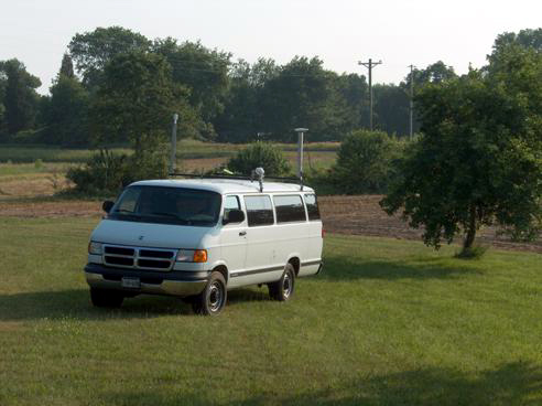 Figure 28. Photo. Van configuration used for multipath testing at the Hagerstown GWEN site. In this case, HA-NDGPS marine antenna to van marine antenna is under test. Picture of a large white van with two antennas mounted on top.  The van is facing toward the lower left hand corner of the photo. On the left side of the van toward the front is a HA-NDGPS antenna; on the right side of the van is mounted the GPS antenna.