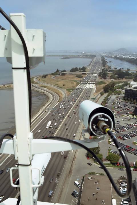 Figure 2. Photograph. A digital video camera mounted on top of a building that overlooks a highway is recording vehicle trajectory data. From atop an elevated mounting frame, a camera looks onto a multilane highway and into the distance toward an interchange.