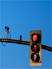 This is a photo of a three-light traffic signal mounted on a black horizontal bar. The traffic signal is red. A traffic camera is also mounted on the black bar to the left of the traffic signal.