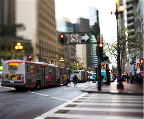 This is a photo of a busy intersection in a city. The photo overlooks a crosswalk with white-striped lines. A line of busses is stopped in the crowded road. The sidewalks are crowded with people.