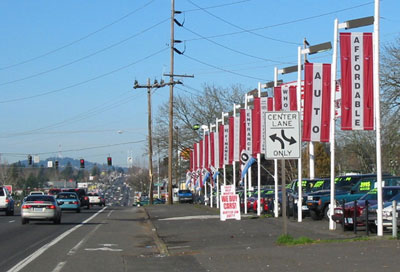 This photo shows an example of sign clutter where the regulatory sign with center lane only arrows is difficult to isolate from the busy background of “affordable,” “entrance,” and “finance” banners and the “we buy cars” sign. 