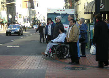 The picture shows a city intersection with a crosswalk delineated with pavers. Seven pedestrians are preparing to cross the street. Six are standing and one is in a wheelchair. 