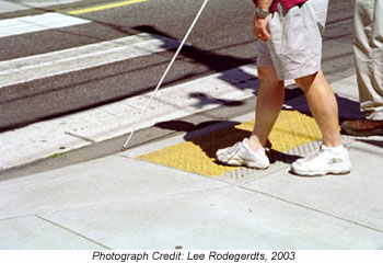 The picture shows a visually impaired person using a cane stepping onto the detectable warning surface of a ramp, about to enter the street. Photograph Credit: Lee Rodegerdts, 2003
