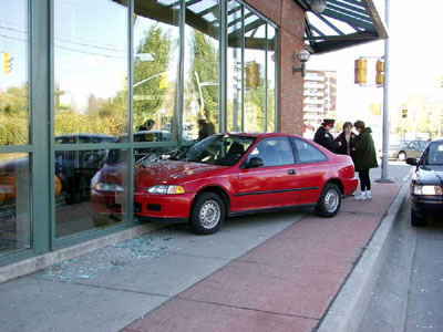 The picture shows a car that has struck a plate-glass window of a building. a police officer is talking with two witnesses in the background on the corner of a signalized intersection. 