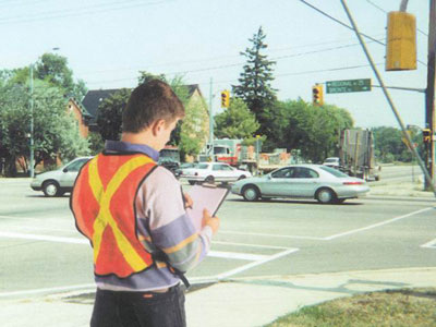 The photo shows a person taking notes and observing traffic conditions at an intersection. The observer is wearing a reflective orange vest. 