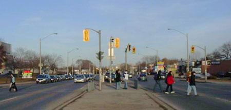 The photo shows a six-lane road with a turn lane and a wide median, with pedestrians in the crosswalks on either side of the median. Pedestrian displays are provided in the median. Fences are provided within the median to discourage pedestrians from crossing in one stage.