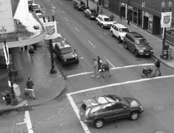 Figure 1. Photo. Many people who use crosswalks are traveling at different speeds. Pedestrian walking speeds generally range between 0.8 to 1.8 meters per second (2.5 to 6.0 feet per second). The picture shows a city street with a crosswalk and pedestrians with different speed characteristics. A man pushes a baby stroller, and three other pedestrians crossing the street on a marked crosswalk. 