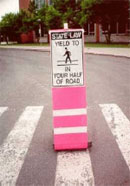 Figure 10. Photo. Regulatory pedestrian crossing sign in New York State. This photo is a close-up of a sign placed in the middle of a crosswalk. The bottom of the sign is orange and white, and the top of the sign reads "State law, yield to pedestrians in your half of the road." The word "pedestrians" is represented by a symbol of a figure walking in a crosswalk.