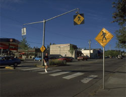 Figure 5. Photo: High visibility crossing with pedestrian crossing signs in Kirkland, WA, The photo sows an intersection on a four-lane road.  This photo shows a lone pedestrian in a crosswalk that is marked by three different yellow and black signs with the crosswalk symbol-one on each side of the street, and one directly over the middle of the crosswalk.