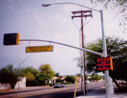 Figure 6. Photo. Experimental pedestrian regulatory sign in Tucson, AZ.  This photo shows a two-lane road. A light post with a yellow sign that reads - pedestrian crossing - stretches over the road, and a sign affixed to post itself reads - stop for pedestrians in crosswalks - bright red letters.
