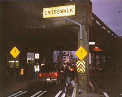 Figure 8. Photo. Overhead crosswalk sign in Seattle, WA.  This photo shows a pedestrian crossing a street at night. The pedestrian is in a crosswalk, which is marked by two different yellow and black signs with the crosswalk symbol on either side of the street, and a sign that reads -crosswalk- hanging over the middle of crosswalk.