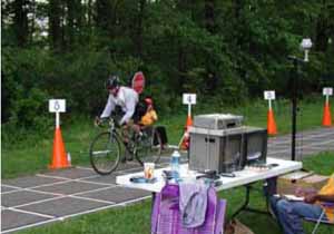 Figure 22: Photo. Video cameras were setup to record participant movements at Stations 3 through 7. A bicyclist is riding through the deceleration station. An event staff person adjacent to the trail is holding up a STOP sign. A video camera is mounted adjacent to the trail and is filming the bicyclist's reaction to the STOP sign.
