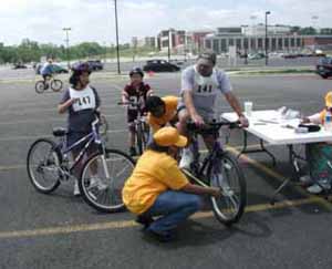 Figure 26: Photos. Physical measurements. Photo 26a: Two event staff persons are measuring the height of a participant in a manual wheelchair. Photo 26b: Two event staff persons are measuring the length of a bicycle and the width of the bicyclist. Two other bicyclists wait their turn.