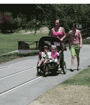 Figure 3: Photo. Jogging stroller for two. A woman is pushing a stroller on a trail. A girl is walking next to her. Two young children are sitting inside the stroller.
