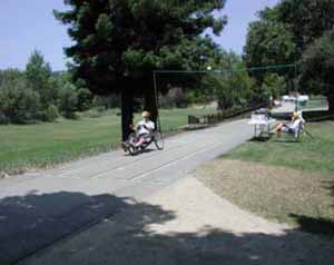 Figure 39: Photo. Speed (and sweep width) station. This is a photo of what actually took place as represented by the drawing in figure 38. We see a section of trail is marked with longitudinal lines spaced 1 meter apart. A participant on a hand cycle is travels through this station. Transverse lines mark the beginning and end of the sweep width and speed station. An overhead video camera is films him. An event staff person is sits at a table and observes the participant.