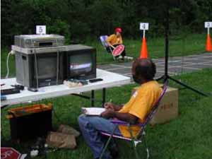 Figure 41: Photo. This is a photo of what actually took place as represented by the drawing in figure 40. Several video cameras were positioned at strategic points around the braking area. This section of trail is marked with longitudinal and transverse lines to form a 1-meter reference grid. An event staff person is sitting alongside the trail and is getting ready to raise a STOP sign. Another event staff person is sitting at a table alongside the trail and is watching the two video monitors. A video camera is mounted nearby.