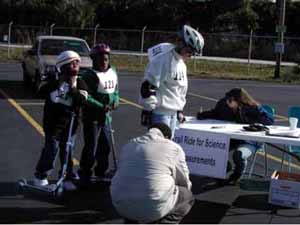 Figure 43: Photo. The study reveals important information on various users now common on shared use paths. An event staff person is taking measurements of a participant using inline skates. Two children on nonmotorized kick scooters wait nearby. Another event staff person is sitting at a table and writing down the measurements.