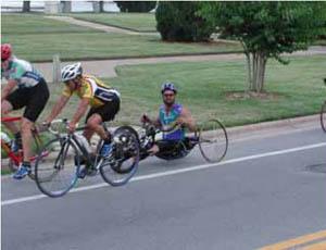 Figure 49: Photo. A hand cyclist. A man is riding a hand cycle in a bicycle lane. Two other bicyclists ride just in front of him.